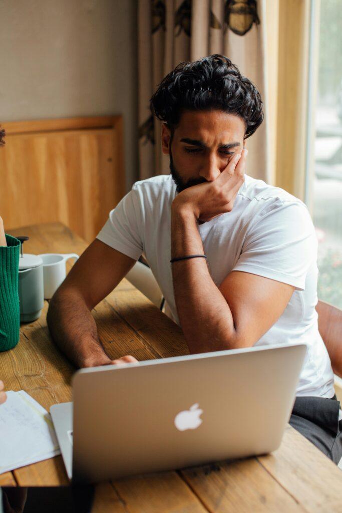 Man in white shirt pondering work on laptop at a cozy indoor setting.