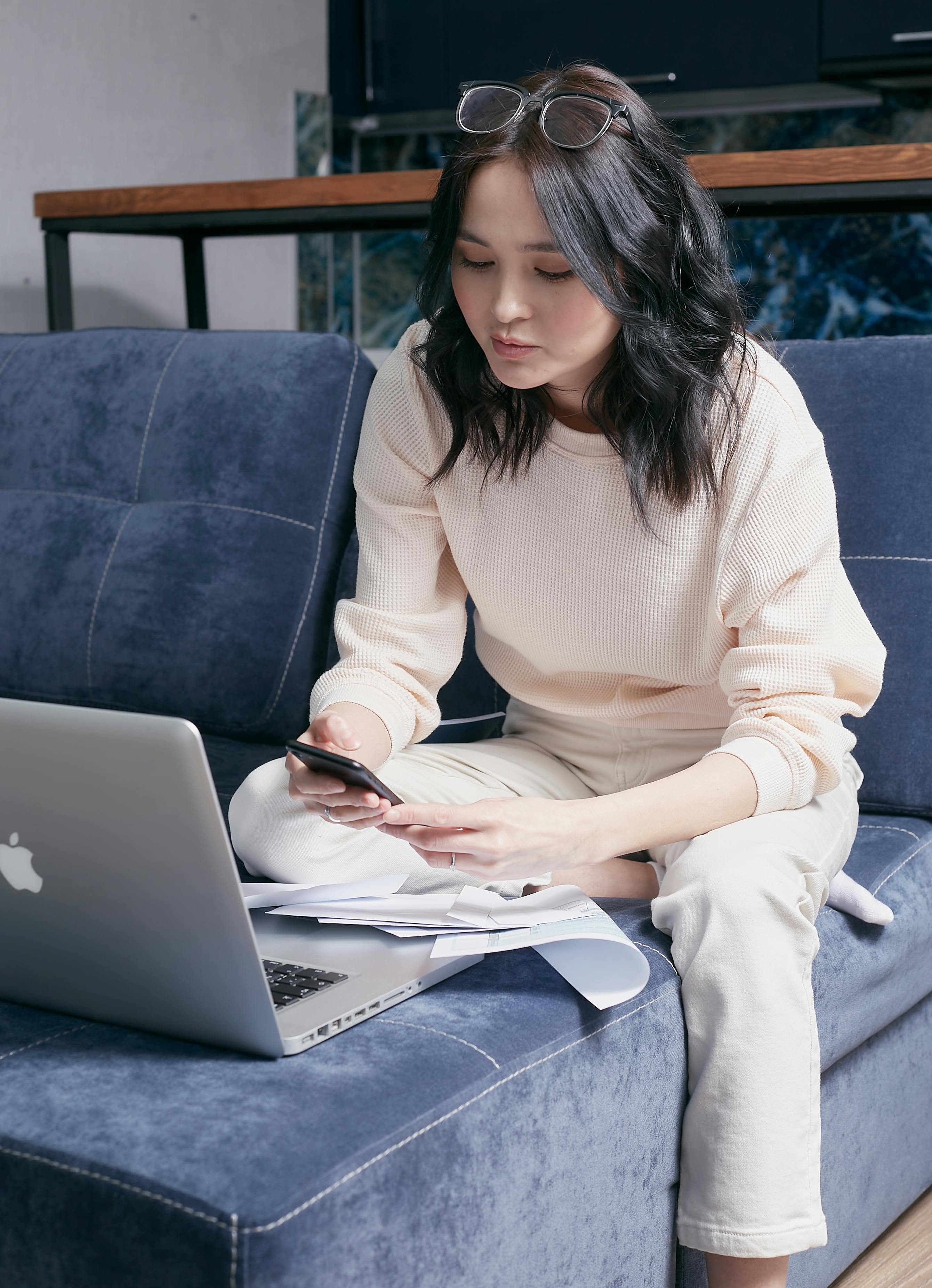 A young woman sits on a sofa managing her bills with a laptop and smartphone.